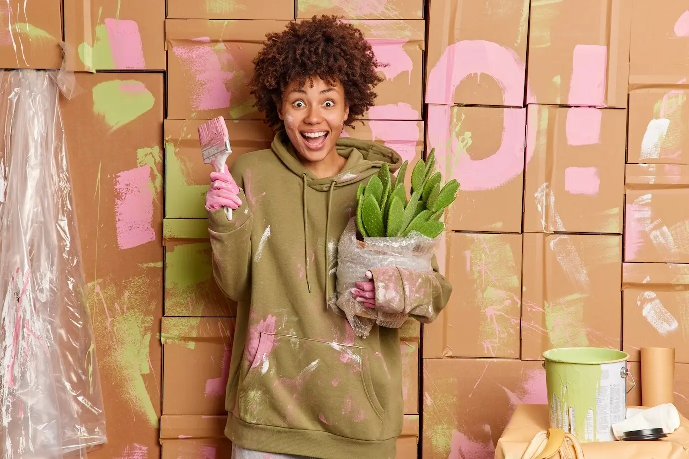 Positiv gestimmte, überglückliche, schmutzige Frau hält einen Kaktus im Topf und einen Pinsel, trägt nach dem Streichen der Wände verschmutzte Kleidung und befindet sich in einem Raum, der von Farbeimern umgeben ist; Konzept: Menschen, Renovierung und Heimverbesserung.